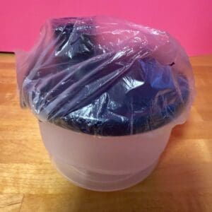A white container with a blue lid wrapped in clear plastic on a wooden table.