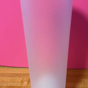 A tall frosted plastic cup on a wooden surface with a pink background.