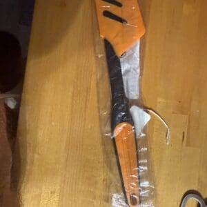 A long knife with an orange and black handle wrapped in plastic on a wooden surface.