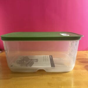 Transparent plastic container with a green lid on a wooden surface against a pink background.