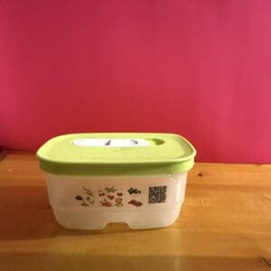 A white container with a green lid sits on a wooden surface against a red wall.