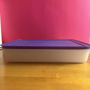 White storage container with a purple lid on a wooden table against a pink wall.