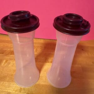Two clear plastic containers with dark lids on a wooden surface against a pink background.